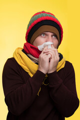 Young sad man wearing burgundy sweater yellow scarf hat hold napkin tissue blowing nose sneeze isolated on plain yellow background studio portrait