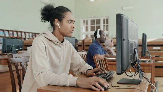 Selective focus shot of young Black student wearing wireless earphones working on desktop computer in college library