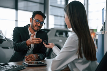 Woman is consulting man in the car showroom. Taking keys from the new automobile