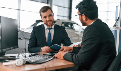 Sitting by the table. Two businessmen are working together in the car showroom