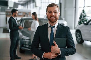 Beautiful man is standing and smiling. Three people are working together in the car showroom