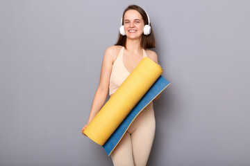Portrait to athletic woman wearing beige sportswear holding yoga mat isolated over gray background, enjoying music in headphones and training in fitness club.