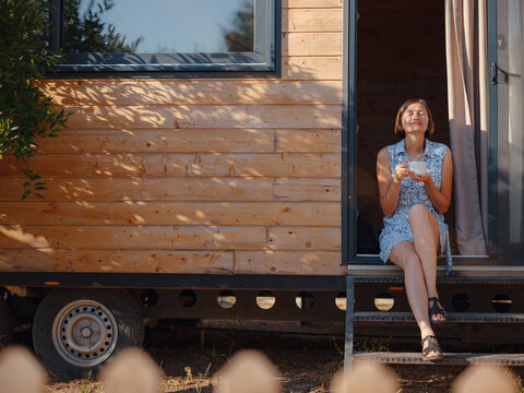 Woman is drinking coffee , morning routine near her Tiny house. First property. Small apartment in summer garden. Minimalism. Moving in. Living alone. Charming trailer house