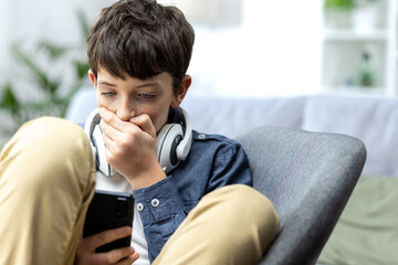 Young teenager boy reading bad news on phone, son sitting on sofa in living room with headphones at...