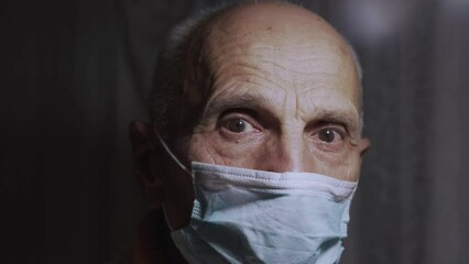 Senior man wearing blue medical mask looks at bright light with attentive and serious expression. Old man looks around spacious dark room