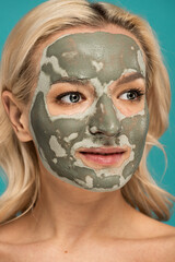portrait of blonde woman with clay mask on face looking away isolated on turquoise.