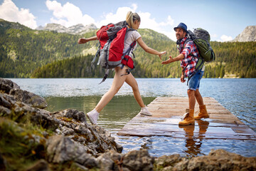 Young couple on wooden jetty by lake. Woman jumping, man lending a hand to her. Summer trip in nature.  Lifestyle, togetherness, nature concept - 587280021