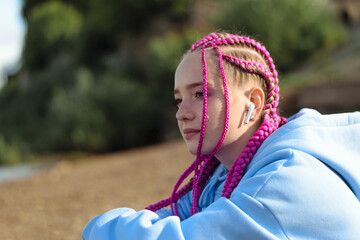 Portrait of a caucasian teenage girl with pink braids using wireless headphones on the river bank.Technology,summer concept.Generation Z style.Copy space.