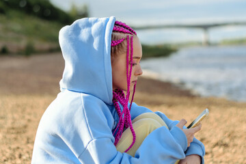 Portrait of a caucasian teenage girl with pink braids using smartphone on the river bank.Technology,summer concept.Generation Z style.
