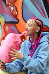 Caucasian teenage hipster girl with pink braids eats cotton candy against the background of a multicolored street wall.Summer concept.Generation Z style.