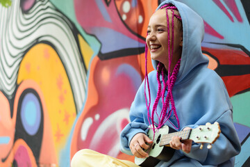 Smiling caucasian teenage hipster girl with pink braids plays the ukulele against the background of a multicolored street wall.Summer concept.Generation Z style.