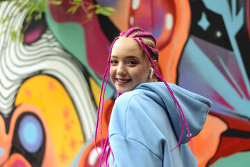 Portrait of a happy caucasian teenage girl with pink braids using wireless headphones against the background of a multicolored street wall.Generation Z style,creativity.
