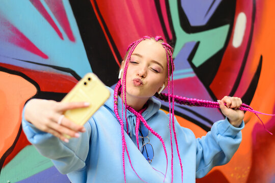Caucasian teenage hipster girl with pink braids is using a smartphone against the background of a multicolored street wall.Summer concept.Generation Z style.Social media concept.