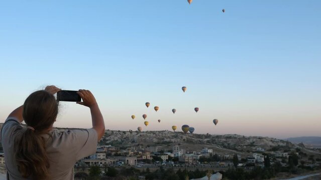 A girl using a smartphone takes pictures of hot air balloons in the sky over Cappadocia. Back view. The most famous place in Turkey
