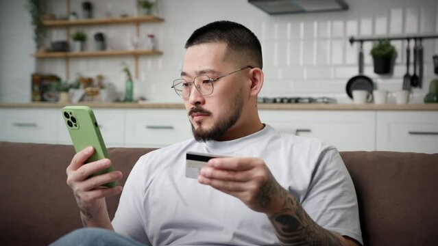 Man Paying Online Using Smartphone. Korean 30s man using bank credit card for online shopping and purchase sitting on couch at home. Close Up.