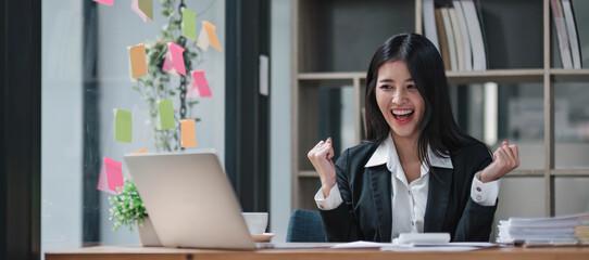 Happy young creative Asian woman working office desk feeling excitement raising her arms up as victory sign, celebrates career ladder promotion or reward. Great results successful work concept © wichayada