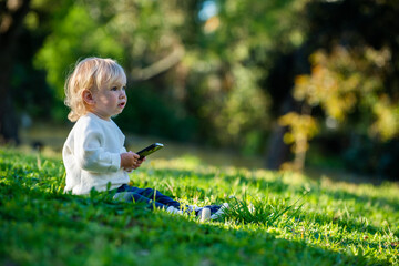 Naklejka na ściany i meble Kid is distracted from his phone while outdoors. A Caucasian child staring away from a smartphone with interest, sitting in a park alone on a sunny day. Cute child sitting in a park on a lovely day.
