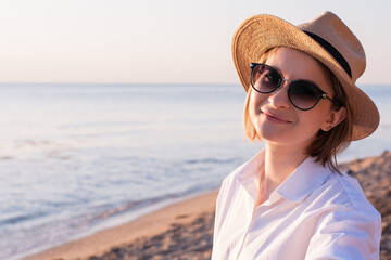 Young adult woman 30 years of mixed race millennial tourist in white shirt straw hat sunglasses taking selfies on the beach on vacation summer at sunset alone while walking outdoors 