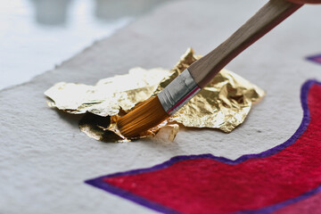 A close up of an artist buffing away gold leaf sheets with brush from artwork to reveal circle sun...