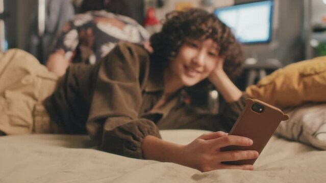Tight medium shot of curly young girl lying in bed in casual clothes, typing on phone, smiling while unrecognizble man sitting behind, turning around, leaving. Turned-on monitor in background, blurred