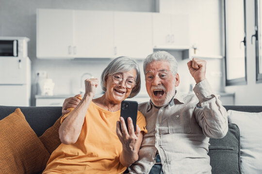 Happy Older Couple Sit On Couch Staring At Cellphone Screen Gesturing Looking Overjoyed, Scream With Joy, Read Fantastic News, Get Great Commercial Offer, Pension Raise. Success, Achievement Concept.