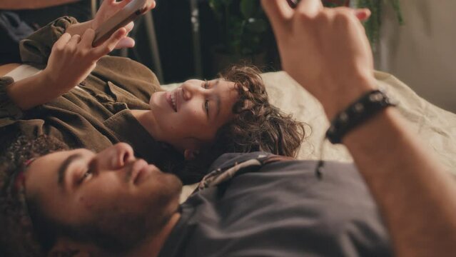 Close-up of young man and woman lying in bed with their phones, head to head, talking, smiling. Sunlight, green plant in background, tilt down shot