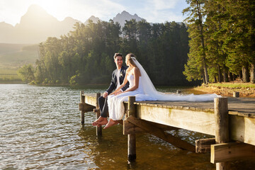 Forest, lake and a married couple on a pier in celebration together after a wedding ceremony of tradition. Marriage, love or romance with a bride and groom sitting outdoor while bonding in nature