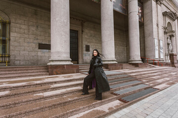 Street photography. A beautiful stylish girl dressed in all black, wearing a long leather coat or jacket, is climbing the steps of a large building with columns.