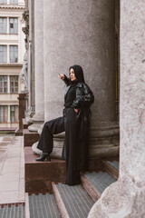 Fototapeta na wymiar Street photography. A beautiful stylish girl dressed in all black, wearing a long leather coat or jacket, is posing against of large building columns.