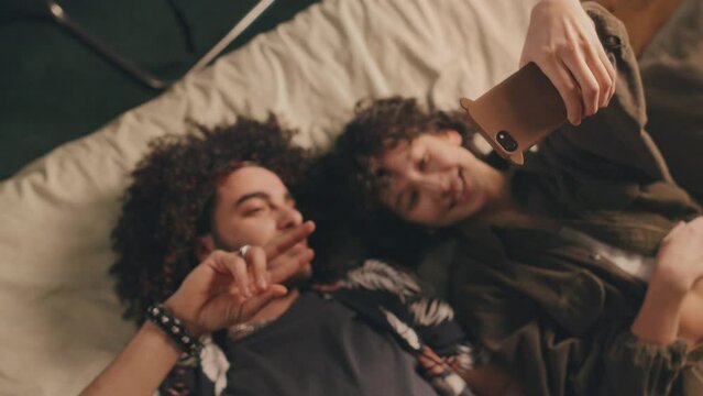 Top down view of curly young couple lying in bed, making V-signs while girl taking selfie of them, both smiling, then showing him pictures, laughing, enjoying themselves