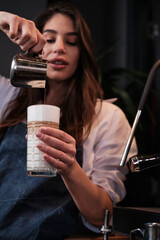 A happy barista preparing fresh coffee and adding milk into a glass while standing next to a coffee machine.