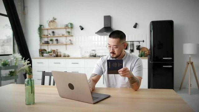 Man Typing on Laptop Holding US Passport in Hands. Asian man using documents working online sitting at home.
