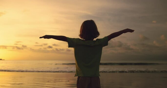 Boy stay at sunset beach spreading hands like bird wings. Kid imagine