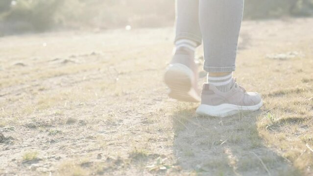 A woman in sports sneakers walks on dried grass and sand. Against the background of the bright rays of the sun, a slow-motion close-up from below.