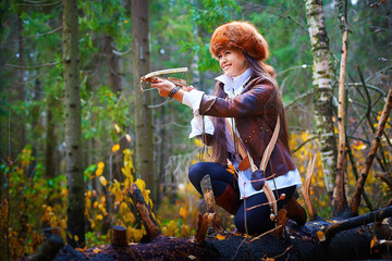 Girl in a leather jacket, a big red fox fur hat and with a crossbow in the forest in autumn. A...