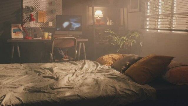 Full medium shot of small cosy bedroom with unmade double bed in foreground, desk with monitor showing video and red lamp in background, all covered with dark, smokey light