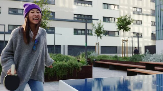 Happy young asian student female having fun playing table tennis with colleague outdoors at college campus. Chinese young millennials enjoying ping pong game on business work break.