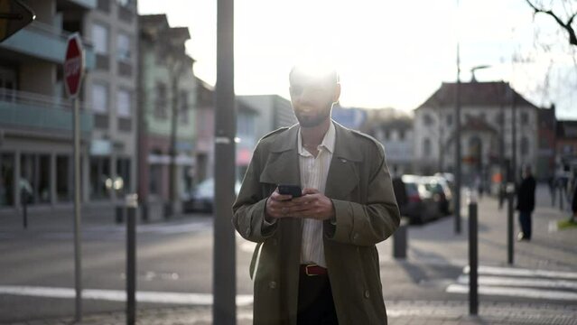 One happy Moroccan young man walking in city street looking at cellphone smiling. A male Middle Eastern person walks in sidewalk during sunset afternoon with flare