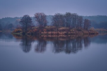 Morning foggy landscape over the water. Trees on an islet on a cloudy day at Lake Mojcza in Kielce, Poland.