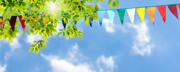 colorful pennant string decoration in green tree foliage on blue sky, summer party background...