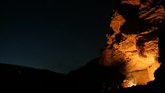 A desert night camping with some people and silhouettes gathering around a bonfire. the fire light is flashing upon the rocks and the skies darken in deep blue. a 4K video clip. Dec 9th, 2022, Israel