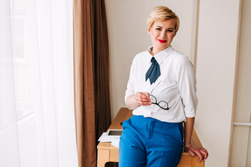 Image of smiling business woman in glasses, in blue costume, company worker, standing over white background near the office table, digital tablet, documents on it