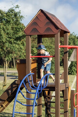A little boy in blue clothes with a hat is playing in a wooden outdoor playhouse