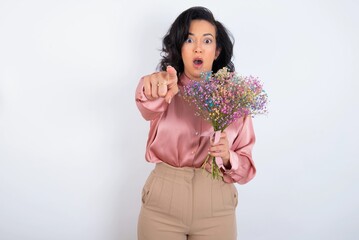 young woman holds big bouquet of nice flowers over white background Pointing with finger surprised ahead, open mouth amazed expression, something on the front.