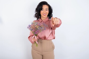 young woman holds big bouquet of nice flowers over white background pointing at camera with a satisfied, confident, friendly smile, choosing you