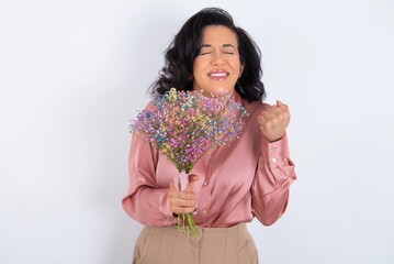 young woman holds big bouquet of nice flowers over white background being excited for success with raised arms and closed eyes celebrating victory. Winner concept.