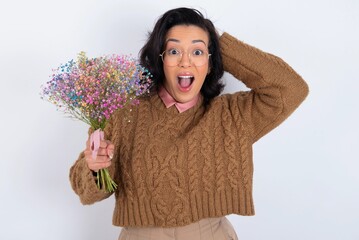 Cheerful overjoyed young woman holds big bouquet of nice flowers over white background reacts rising hands over head after receiving great news.