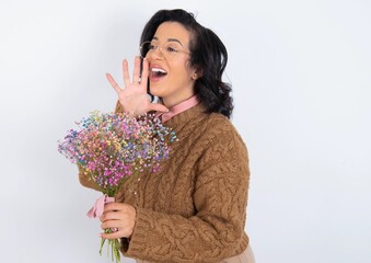 young woman holds big bouquet of nice flowers over white background look empty space holding hand face and screaming or calling someone.