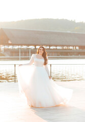 Fototapeta na wymiar A beautiful brunette bride in a white dress is circling the lake at sunset
