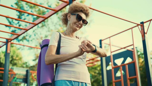 Happy elderly woman in good physical shape uses fitness tracker after yoga class outside. Modern sports gadgets, the concept of fitness equipment. Sports smart watch. Healthy lifestyle on retirement. 